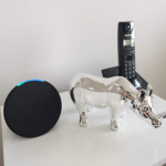 Dispositivo inteligente en forma circular, una figura decorativa plateada de un rinoceronte y un teléfono inalámbrico negro, dispuestos sobre una superficie blanca.