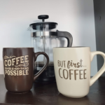 Dos tazas de cerámica con frases relacionadas al café, acompañadas de una prensa francesa de vidrio y plástico negro, colocadas sobre una superficie.
