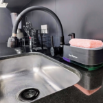 Fregadero de acero inoxidable con grifo moderno de diseño negro mate, acompañado de un dispensador de jabón con esponja rosada y utensilios de cocina organizados al fondo.