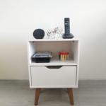 Mesa auxiliar blanca con patas de madera, equipada con un altavoz inteligente, una figura decorativa plateada de un rinoceronte, un teléfono inalámbrico, un contenedor de plástico, un costurero con hilos de colores y un cajón que contiene un router de internet.