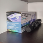Plancha Black+Decker en su caja original con especificaciones visibles y un paraguas negro plegado, ambos colocados sobre un estante de madera.