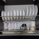 Escurridor de vajilla de acero inoxidable con platos blancos, vasos de vidrio y tazas colocados en un armario de cocina.