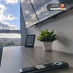 Escritorio moderno con una planta decorativa, un control remoto, un marco de foto y un televisor montado en la pared. Al fondo, una ventana amplia con una vista despejada y cielo azul. Logotipo de Superhost de Airbnb en la esquina superior derecha.