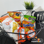 Canasta de bienvenida con snacks variados, incluyendo papas Margaritas, Quinua Pops y Palomitas, decorada con una planta en maceta y un portavasos con servilletas.