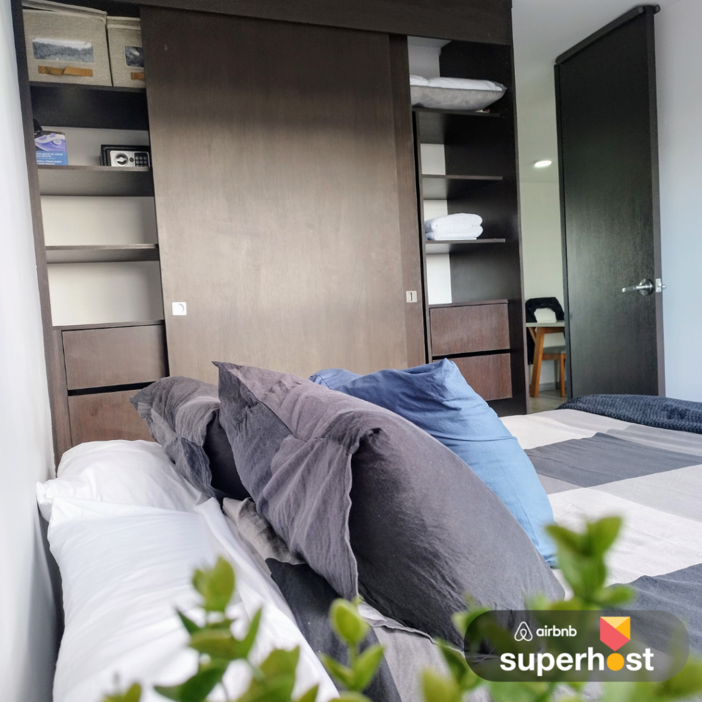 Vista lateral de una cama con cojines grises y azules en un dormitorio moderno, con un armario de madera corredizo al fondo y el logo de Airbnb Superhost en la esquina inferior derecha.