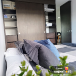 Vista lateral de una cama con cojines grises y azules en un dormitorio moderno, con un armario de madera corredizo al fondo y el logo de Airbnb Superhost en la esquina inferior derecha.