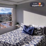 Habitación luminosa con cama doble decorada con ropa de cama azul y blanca, junto a una ventana amplia con vista a la ciudad y montañas. Logotipo de Superhost de Airbnb en la esquina superior derecha.