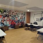 Espacio de coworking y videojuegos con un mural de arte urbano y mesas de trabajo.
