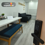 Vista de la entrada al apartamento con una cerradura digital iluminada, un comedor con banco tapizado en azul y una sala con sofá negro. En la pared se aprecian cuadros decorativos y una cortina tipo sheer screen cubriendo la ventana.