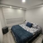 Habitación con cama matrimonial vestida con ropa de cama en tonos azules y blancos, cortinas tipo sheer screen, iluminación LED empotrada y un cesto de ropa en la esquina.