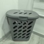 Cesto de ropa sucia de plástico gris con diseño ventilado y tapa, ubicado en una esquina de la habitación cerca de una cortina.