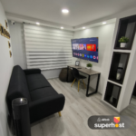Espacio de trabajo con un escritorio de madera, silla negra con patas de madera, televisor Smart TV montado en la pared con acceso a plataformas de streaming, puff de descanso y estanterías empotradas con decoración.