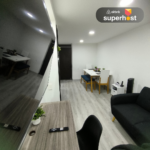 Vista de la sala-comedor con un escritorio de madera, Smart TV, sofá negro, mesa de comedor con sillas y banco tapizado, además de una mesa auxiliar con teléfono y altavoz inteligente.